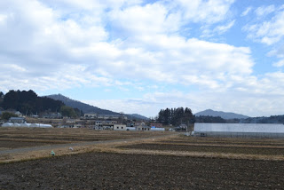 雲が多い青空の下、広々とした田畑の奥にビニールハウスがあり、そのさらに奥に住宅と山並みが見える風景写真