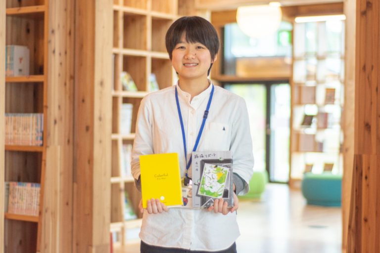 木製の柱や本が並んでいる棚の前に立つ、2冊の本を両手で持った、白いシャツを着た小林さんの写真