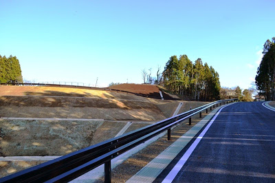 黒く舗装された広い道路がゆるやかにカーブしながら続いており、道路脇の斜面は土が整地された造成地の写真