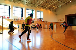 体育館の広いフロアで、ピンクと緑のビブスを着た選手がボールを蹴ってパスを回しているフットサルの試合中の写真