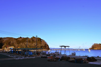 漁港の岸壁沿いに漁具やロープが置かれ、青空の下にいくつかの漁船が浮かぶ静かな海が広がっている港の風景の写真