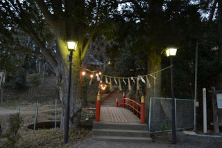 奥には神社へと続く石段が見える両側に大きな木が立ち、灯りが灯された両側の街灯に白い紙垂がついた伝統的な注連縄が掛けられ、赤い欄干と金色の装飾が施された小さな木橋の入り口の写真