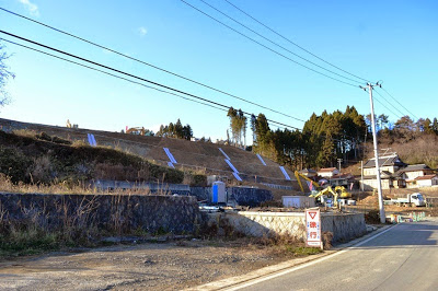 住宅が点在する山のふもとの斜面で、白いシートが貼られた整地作業が進められ、道路沿いに工事用車両や重機が見える宅地造成中の斜面の写真