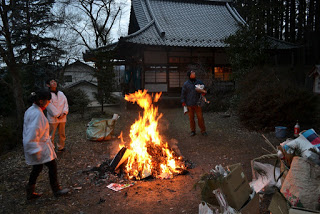 夕暮れ時に八幡宮の境内で持ち寄った正月の松飾りやしめ縄、古神符がお焚き上げされた火を囲む人々の写真
