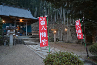 周囲を木々が立ち並ぶ森の中にあり、赤い奉納旗が両側に立ち、石段と灯りが灯された灯籠が並ぶ八幡宮の神社の写真