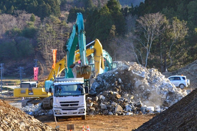 背景には木々に囲まれた山林が広がっている造成工事現場で、複数の重機が大きな岩の山を崩してダンプカーに積み込む作業を行っている写真