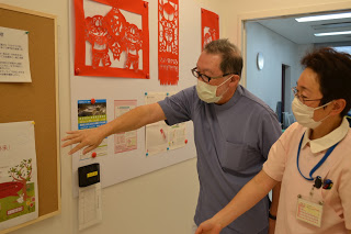 たくさんの文書や切り絵が掲載された、壁の掲示スペースを指しながら話をしている中村先生と齋藤さんの写真