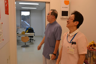 扉を開けた診察室の前で中村先生と、白い襟付きの薄いピンク色のユニフォームを着た笑顔の齋藤さんが立っている写真