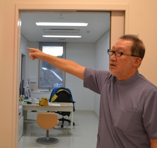 机の両側に椅子が配置された診察室の前で、右側を指さして話をしているグレーのユニフォームを着た中村幸夫先生の写真