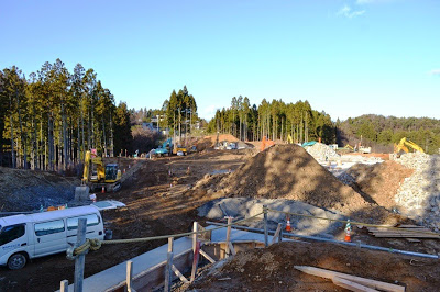 複数の重機が並ぶ林に囲まれた広い造成地で、土砂の山や杭が打たれた基礎部分が確認できる造成工事中の住宅地予定地の写真