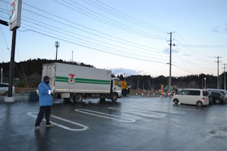 乗用車やセブンイレブンの大型トラックが停まっている駐車場に、青い防寒着の男性が立っている写真