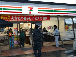 オレンジと緑、赤の配色のセブンイレブンのマークとストライプ模様がデザインされた店舗の中と外が訪れた人々でにぎわっている様子の写真