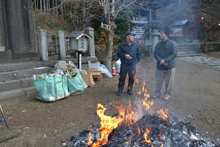 背景に木々が見える八幡宮の境内に、しめ縄などが入った紙袋があり、お焚き上げの火のそばに立つ二人の人物が並んで立ち話をしている様子の写真