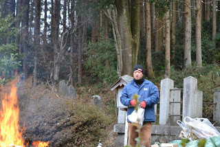 石碑が並ぶ八幡宮の境内で、青い上着と赤い手袋を着けた男性が、お焚き上げされた火のそばで袋に入った松飾りを持って見ている様子の写真