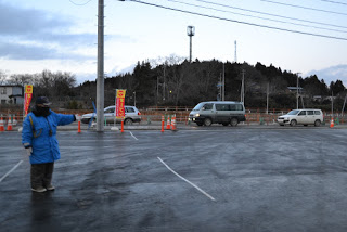 車が連なって走る道路横の駐車場で、青い防寒着の男性が車の誘導を行っている写真