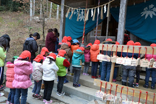 右側に絵馬が掛けられ、背景に白い模様の青い幕が垂れた社殿の前に、赤い帽子とカラフルな上着を着た子どもたちが並んで手を合わせて拝んでいる様子の写真