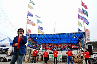 屋根が青色の舞台が奥にあり、その前に赤いジャンバーを着た関係者が5名並び、1番前でマイクを持ち歌を歌っている亜KIRAさんの写真