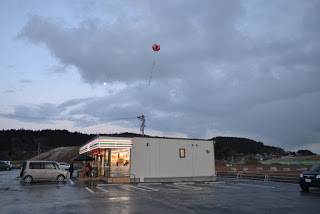 薄暗い中、広い駐車場に囲まれたセブンイレブンの店舗の灯りが煌々と輝き、店舗の上には赤いアドバルーンが浮かんでいる写真