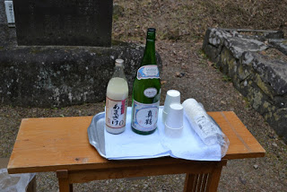 石碑の前に設置された木のテーブルの上に緑と白の酒瓶、白い紙コップが載った盆が置かれている様子の写真