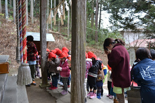 しめ縄が飾られた八幡宮の社殿の前に先生に付き添われ、赤い帽子をかぶった子どもたちが社殿の前に整列し手を合わせて拝んでいる様子の写真
