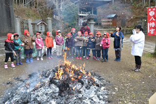 木々に囲まれた八幡宮の境内で、右側に2人の女性が立ち、赤い帽子とカラフルな上着を着た子どもたちがお焚き上げされた大きな焚き火を囲んでいる様子の写真