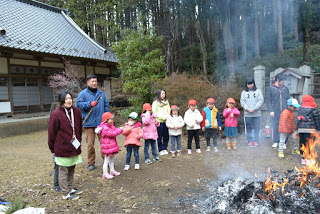 木々に囲まれた八幡宮の境内で、色とりどりの冬服を着た子どもたちと先生方がお焚き上げされた小さな焚き火を囲んでいる様子の写真