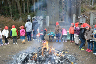 大きな石碑のような構造物がある八幡宮の境内で、赤い帽子をかぶった子どもたちがお焚き上げされた焚き火の前に立っている様子の写真