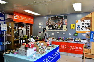 テーブルに沢山の土産品が陳列されている、オレンジ色や青色の「南三陸さんさん商店」と書かれた幕が飾られた店舗内を、2人の買い物客が商品を見ている写真