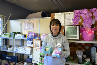 線香やろうそくなどが整然と並んだ南三陸お土産ショップの一角で、女性スタッフが笑顔で商品を両手に持って紹介している写真