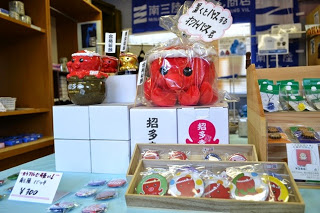 店内の棚に、赤いタコの形をしたオクトパス君のぬいぐるみや缶バッジなどのグッズが整然と並べられている写真