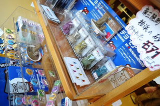 南三陸限定のモアイ型の置物や缶バッジが陳列棚に並び、手書きの値札が飾られている写真