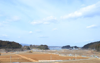整地された広大な土地が広がり、遠くに海と山々が見え、手前には工事車両や作業現場が点在している写真