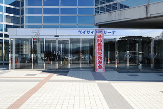 ガラス張りの近代的な建物の正面入口に「高齢者芸能発表会」と書かれた縦長の看板が立てられており、上部には「ベイサイドアリーナ」の文字が掲げられている、伝統芸能発表会の会場入口の写真