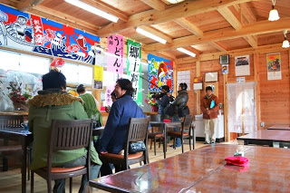 木組みの内装で大漁旗が掲げられている温かみのある店内で、地元客と思われる数名がカウンターやテーブル席に座り、リラックスして食事を楽しんでいる郷土料理店の内部の写真