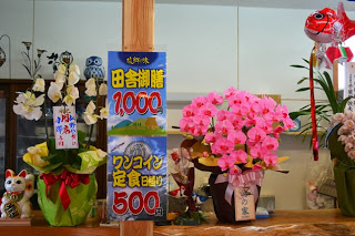 カウンターに並んだ豪華な鉢植えの花とともに「田舎御膳1,000円」「ワンコイン定食500円」といったメニュー案内が掲示されている様子の写真