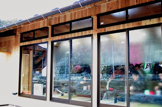 ガラス窓越しに店内を外から見た様子で、多くのお客さんがテーブル席に座り、にぎやかに食事や会話を楽しんでいる郷土料理店の写真。