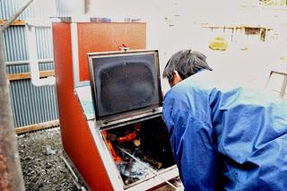 青い作業着の男性が、屋外に設置された薪を燃料とする大型ボイラーの蓋を開け、中を確認している様子を写した写真