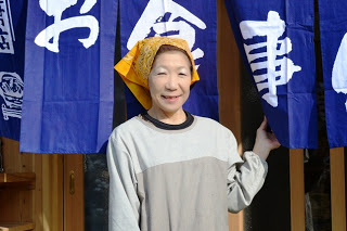暖簾を前に笑顔で写る松野三枝子さんの写真