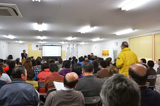 前方にスクリーンが設置され、講師の男性が壇上で話をしており、多くの参加者が席について講座に参加している会場の全体の様子を写した写真