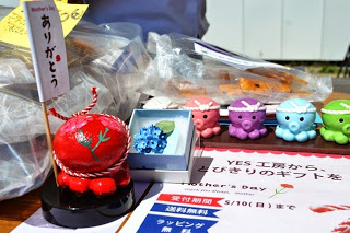 「ありがとう」と書かれた旗のついた赤いオクトパス君の置物や、水色の箱に入った紫陽花の花のブローチ、カラフルな小さなタコの置物などが並び、母の日ギフトの受付期間などが記載されたチラシが置かれているコーナーの写真