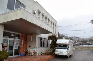 白い壁の建物の前にキャンピングカーが停まり、集会所の入口が見える入谷公民館の外観の写真