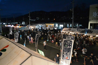 夜の福興市会場に多くの屋台と観客が集まり、夏祭りの熱気に包まれてにぎわっている様子を高い位置から撮影した写真