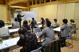 木造の室内でホワイトボードを使って講義を行う男性と、それを真剣に聞く受講者たちの様子を左後方から撮影した写真