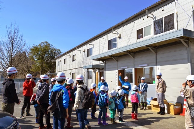 子供たちと大人たちが白いヘルメットをかぶり、建物の前に集まって前方の右手を挙げた男性の話を聞いている写真