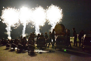 ステージ上で太鼓の演奏が続く中、背景で複数の花火が同時に打ち上がり、夜空を照らしている写真