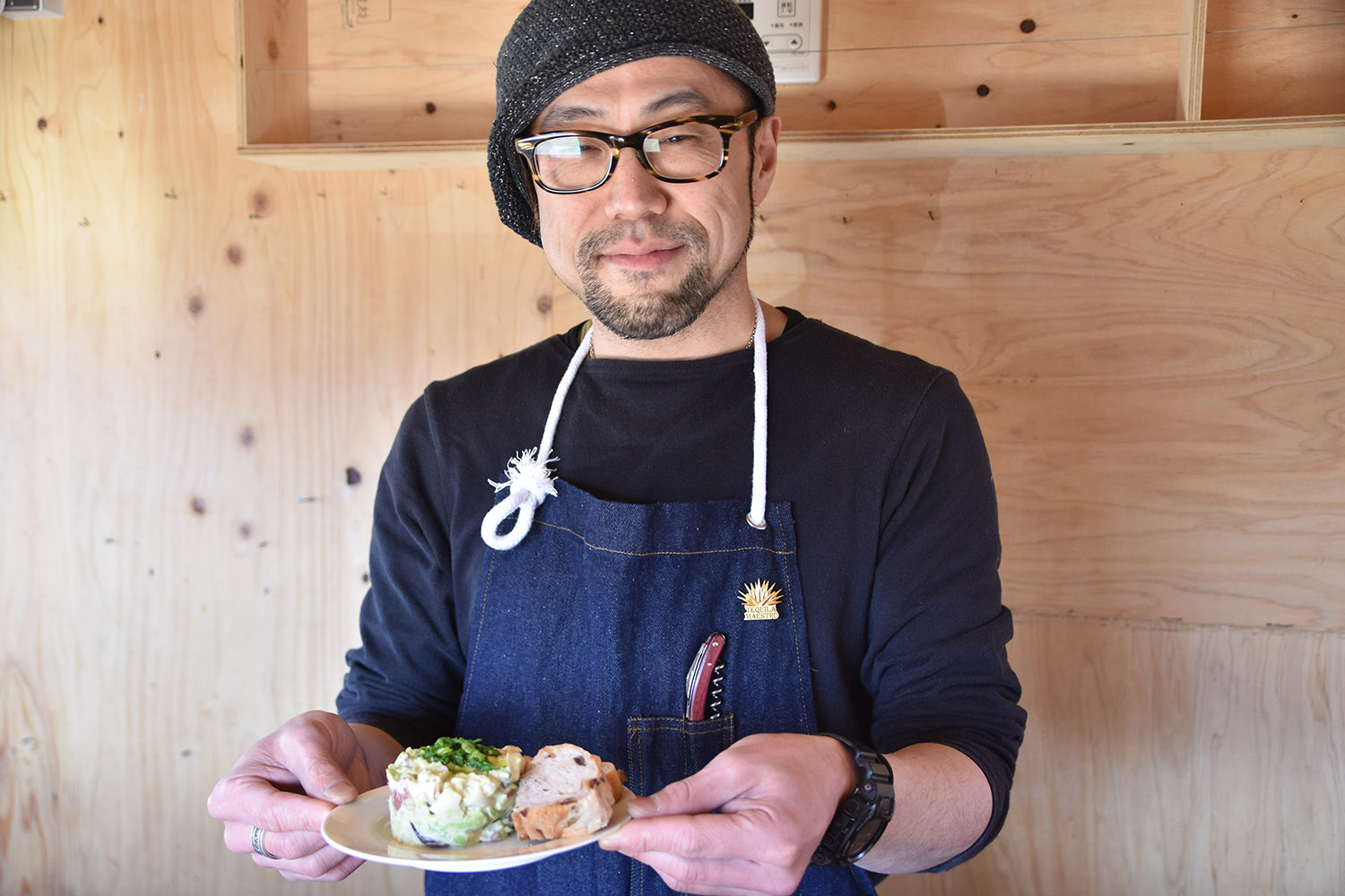 紺色のシャツとデニムのエプロンを着た男性が、木製の壁の前で小さな皿に盛られた2つの料理を手に持っている様子を写した写真