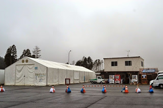 手前にカラーコーンが並べられている舗装された広い駐車場の奥に、白い大型テントとその隣に建つ二階建ての建物が並んでいる写真