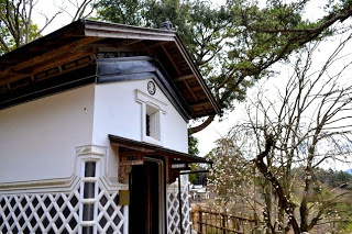 白い壁と黒い屋根を持ち、壁には格子模様が施され、建物の上部には小さな窓があり、その下に木製の扉がある建物の写真