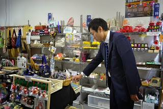 棚に並べられた様々なお菓子や小物、袋詰めの商品などが陳列され、商品を見ているスーツ姿の男性が右手でバスケットの中の商品を指差している写真