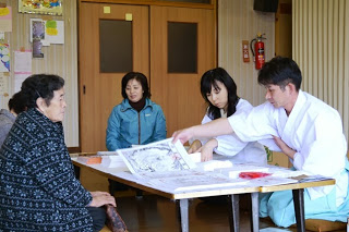 神職が高齢の女性に丁寧に説明し、他の住民がその様子を見守っている室内の写真